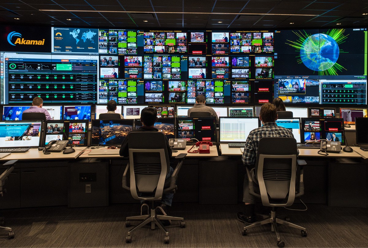 Six people working in a Broadcast Operations Control Center with video walls and consoles